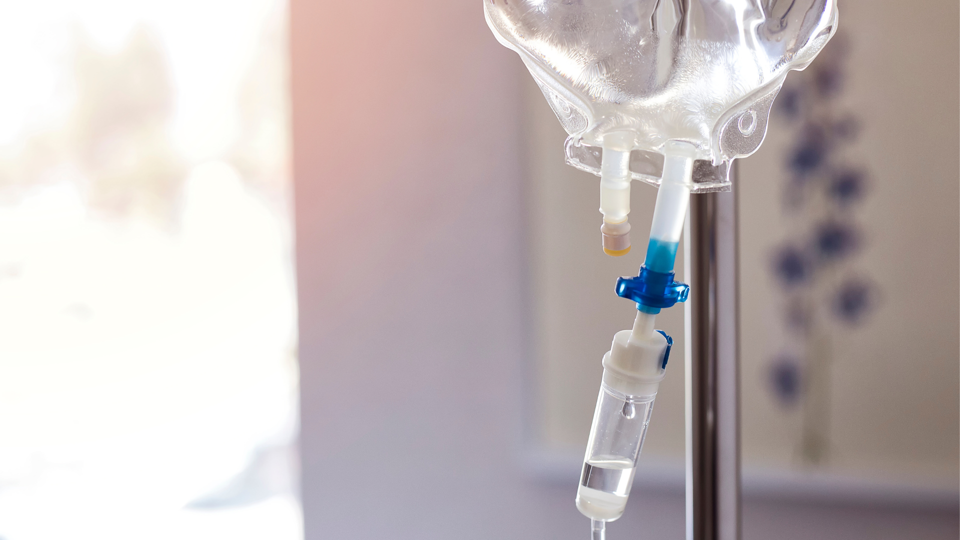 A clear IV fluid bag and blue drip line hanging on a stand in a bright treatment room